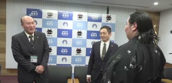 相撲甚句で祝福する奄美市・向美芳教育長（左）。中央は安田壮平市長