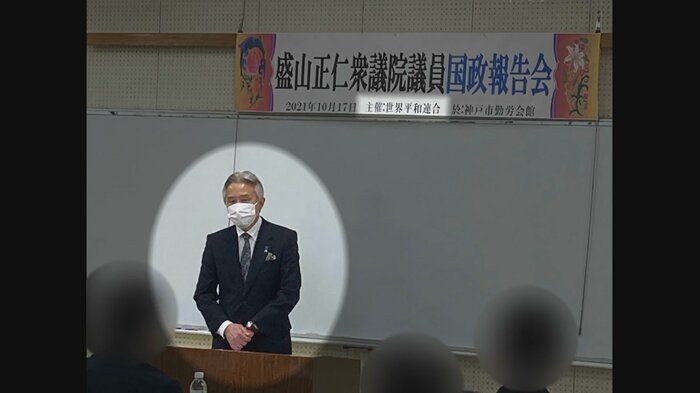 「世界平和連合」と書かれた看板を背に挨拶する盛山氏