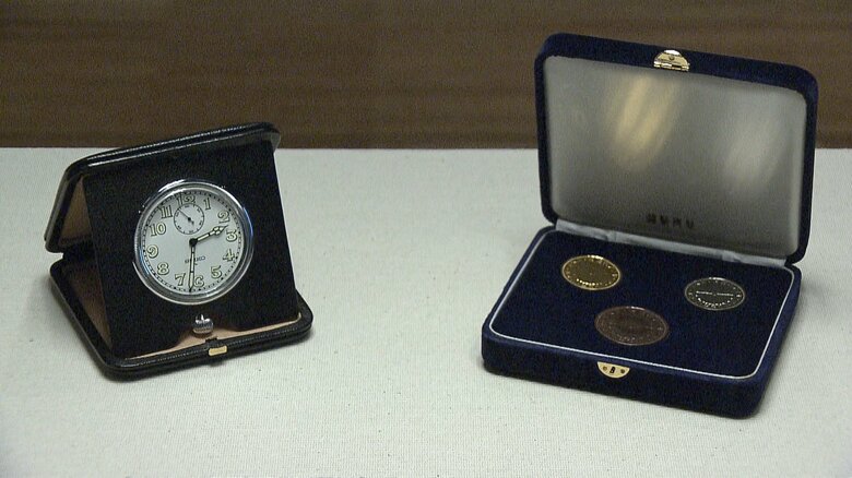 左側：枕時計　服部時計店（昭和40年代）、右側：昭和天皇在位60年記念貨幣　大蔵省造幣局（1986年）皇居三の丸尚蔵館所蔵