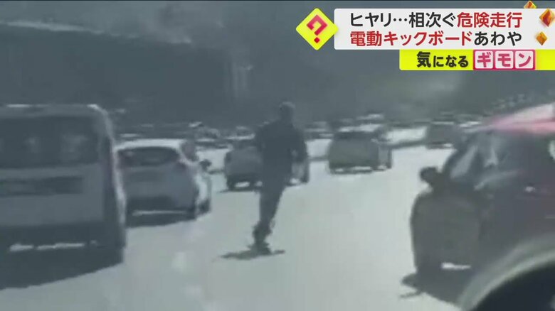 スピード違反をしながら車の横をすり抜け走り去っていくキックボードの男性
