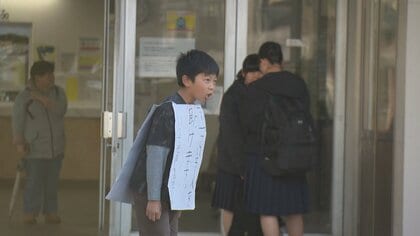 「困っている人を助けます」の手作り看板をぶら下げて駅に立つ小学生 「人助け」の活動を続ける理由
