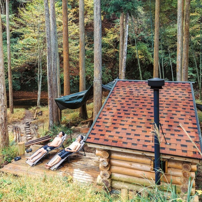 スギ林に囲まれた丸太のサウナ小屋とウッドデッキ。奥には川が流れ、サウナを楽しむには絶好の環境(『小屋を建てる』より)